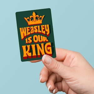 Pode incluir: Um autocolante retangular verde com o texto "WEASLEY IS OUR KING" em laranja e vermelho, encimado por uma coroa dourada. O autocolante é segurado por uma mão contra um fundo azul claro.