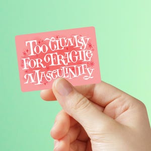 May include: A pink rectangular sticker with the text "Too Clumsy For Fragile Masculinity" in white script. The sticker is held by a hand against a light green background.