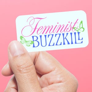 May include: A rectangular sticker with the words "Feminist BUZZKIL" in pink and blue, with butterfly graphics. The sticker is held against a pink background.