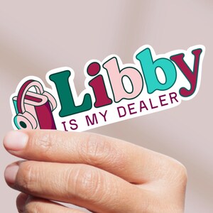 May include: A colorful sticker with the text "Libby IS MY DEALER" in a playful font. The sticker features a teal, pink, and burgundy color scheme, with a graphic of a yoga mat and headphones. The sticker is held by a hand.