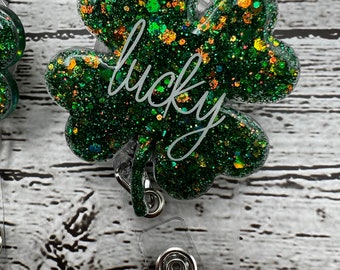 May include: A green glitter shamrock badge reel with the word "lucky" in white script. The badge reel has a clear plastic strap and a metal clip for attaching to clothing. The shamrock is a symbol of good luck.