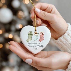 May include: A heart-shaped Christmas ornament with a gold string, featuring three stockings in green, blue, and red. The ornament reads "FIRST CHRISTMAS AS Mommy & Daddy 2025". The stockings are decorated with festive details, including a teddy bear.