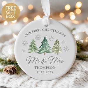 May include: White ceramic Christmas ornament with a ribbon for hanging. The ornament features watercolor illustrations of pine trees and snowflakes, with the text "OUR FIRST CHRISTMAS AS Mr & Mrs Thompson 11.19.2025".