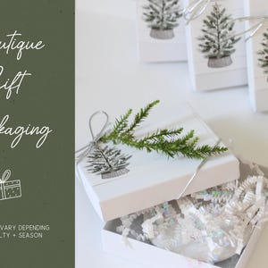 May include: White gift boxes with a Christmas tree design and silver ribbon. One box is open, revealing white shredded paper and a sprig of greenery. The text "Boutique Gift Packaging" is visible on the left side of the image.