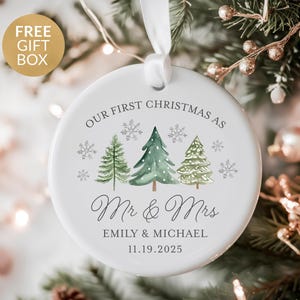 May include: White ceramic Christmas ornament with watercolor trees and snowflakes. Text reads "OUR FIRST CHRISTMAS AS Mr & Mrs EMILY & MICHAEL 11.19.2025". Includes a white ribbon for hanging. A gold gift box is in the upper left corner.