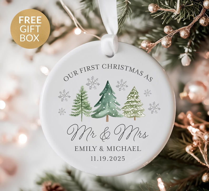 May include: White ceramic Christmas ornament with watercolor trees and snowflakes. Text reads "OUR FIRST CHRISTMAS AS Mr & Mrs EMILY & MICHAEL 11.19.2025". Includes a white ribbon for hanging. A gold gift box is in the upper left corner.