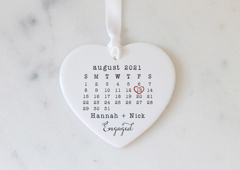 K&ouml;nnte beinhalten: Wei&szlig;er, herzf&ouml;rmiger Anh&auml;nger aus Keramik mit einem Kalender f&uuml;r August 2021, wobei der 13. rot umkreist ist. Der Text "Hannah + Nick Engaged" ist unter dem Kalender gedruckt.