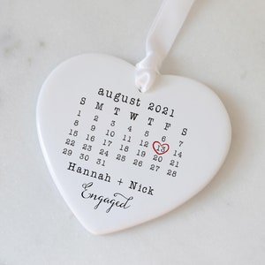 K&ouml;nnte beinhalten: Wei&szlig;er, herzf&ouml;rmiger Anh&auml;nger aus Keramik mit einem Kalender f&uuml;r August 2021. Der 13. ist mit einem roten Herzen umkreist und der Text "Hannah + Nick Engaged" befindet sich unter dem Kalender.