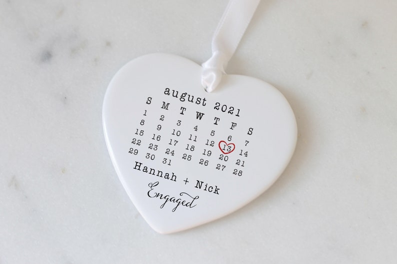 K&ouml;nnte beinhalten: Wei&szlig;er, herzf&ouml;rmiger Anh&auml;nger aus Keramik mit einem Kalender f&uuml;r August 2021. Der 13. ist mit einem roten Herzen umkreist und der Text "Hannah + Nick Engaged" befindet sich unter dem Kalender.