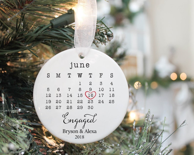May include: White ceramic ornament with a calendar for June with a red heart on the 16th. The text "Engaged" is written in cursive with the names Bryson & Alexa and the year 2018 below.