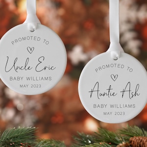 May include: Two white ceramic Christmas ornaments with white ribbon hangers. One reads "Promoted to Uncle Eric, Baby Williams, May 2023." The other reads "Promoted to Auntie Ash, Baby Williams, May 2023." Both have a heart symbol.