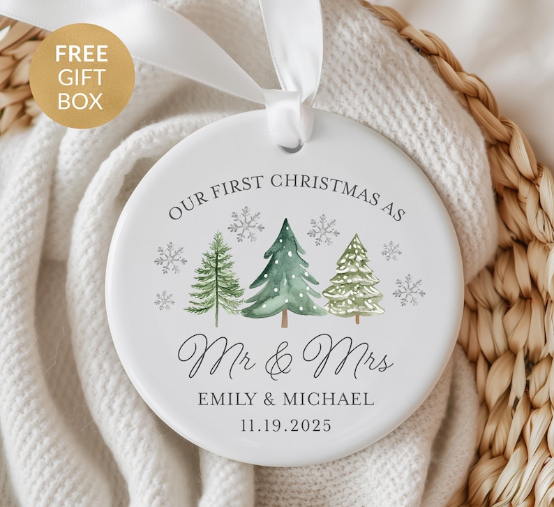 May include: A white ceramic Christmas ornament with a white ribbon. The ornament features the text "OUR FIRST CHRISTMAS AS Mr & Mrs" with the names "EMILY & MICHAEL" and the date "11.19.2025". The design includes watercolor-style Christmas trees and snowflakes.