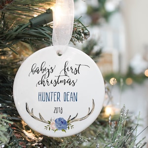 May include: White ceramic Christmas ornament with the text "baby's first christmas" and "HUNTER DEAN 2018". The ornament is decorated with antlers, blue and pink flowers, and green leaves.