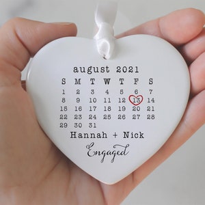 K&ouml;nnte beinhalten: Wei&szlig;er Keramik-Herzen-Anh&auml;nger mit einem Kalender f&uuml;r August 2021. Der 13. ist mit einem roten Herzen hervorgehoben. Der Text "Hannah + Nick Engaged" ist unter dem Kalender gedruckt.