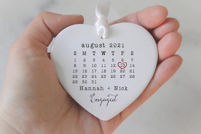 K&ouml;nnte beinhalten: Wei&szlig;er Keramik-Herzen-Anh&auml;nger mit einem Kalender f&uuml;r August 2021. Der 13. ist mit einem roten Herzen hervorgehoben. Der Text "Hannah + Nick Engaged" ist unter dem Kalender gedruckt.