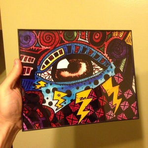 May include: A framed artwork featuring a colorful, abstract eye with a black and white checkered background. The eye is surrounded by swirls, dots, and lightning bolts. The artwork is signed "ZM" in the bottom right corner.