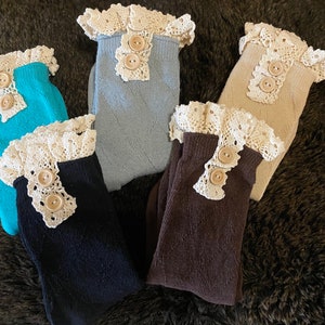 May include: Five pairs of socks in various colors: teal, light blue, beige, navy, and brown. Each sock features a white lace trim with two wooden buttons. The socks are arranged on a dark brown, textured surface.