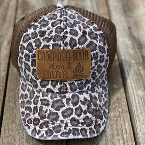 May include: A brown and black leopard print trucker hat with a brown leather patch that says "Camping Hair don't care" with a campfire icon.