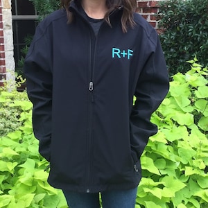 Op de afbeelding: Een zwarte softshelljas met een ritssluiting en een blauw geborduurd logo met de tekst "R+F".