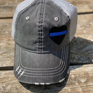 May include: A gray and black baseball cap with a mesh back. The cap features a black heart with a blue stripe and white stitching. The brim has distressed detailing.