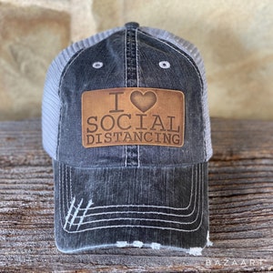 Peut inclure: Un chapeau de camionneur en toile noire et grise usée avec un patch en cuir marron qui dit "I ♥ SOCIAL DISTANCING".