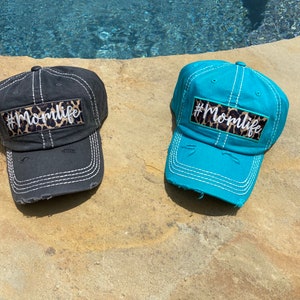 May include: Two distressed baseball caps with a leopard print patch and the text "#Momlife" in white. One cap is black and the other is turquoise.
