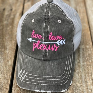 May include: A black and gray distressed trucker hat with a white mesh back. The hat has pink embroidery that reads "live love plexus" with an arrow pointing to the right.