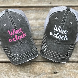 Puede incluir: Dos gorras de camionero grises y blancas con el texto "Wine o'clock" bordado en la parte delantera. Una gorra tiene letras rosas y la otra letras blancas.