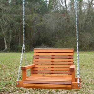 Puede incluir: Un columpio de madera con respaldo y asiento de listones, colgado de dos cadenas plateadas. El columpio est&aacute; en una zona de hierba con hojas ca&iacute;das.
