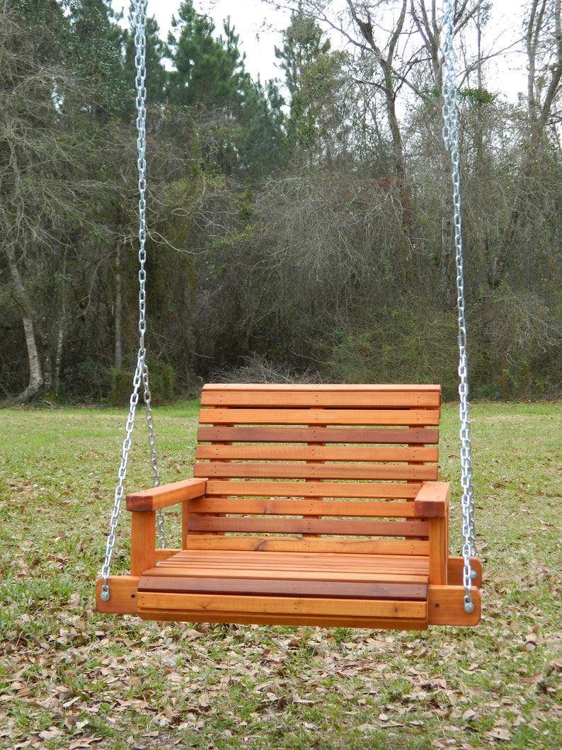 Puede incluir: Un columpio de madera con respaldo y asiento de listones, colgado de dos cadenas plateadas. El columpio est&aacute; en una zona de hierba con hojas ca&iacute;das.