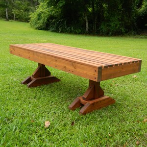 May include: A long wooden bench with a natural brown stain finish. The bench has a slatted top and is supported by two sets of angled legs.
