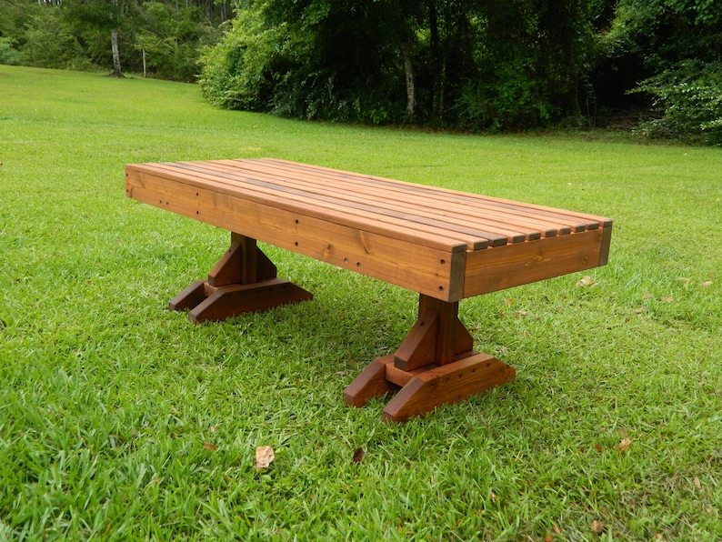 May include: A long wooden bench with a natural brown stain finish. The bench has a slatted top and is supported by two sets of angled legs.