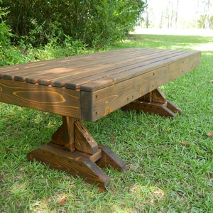 May include: A dark brown wooden bench with a slatted top and a unique, sturdy base. The bench is sitting on a grassy lawn.