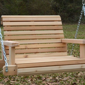 Puede incluir: Un columpio de madera con respaldo de listones y apoyabrazos, colgado de dos cadenas plateadas. El columpio est&aacute; hecho de madera de cedro natural y est&aacute; suspendido en un &aacute;rea cubierta de hierba.