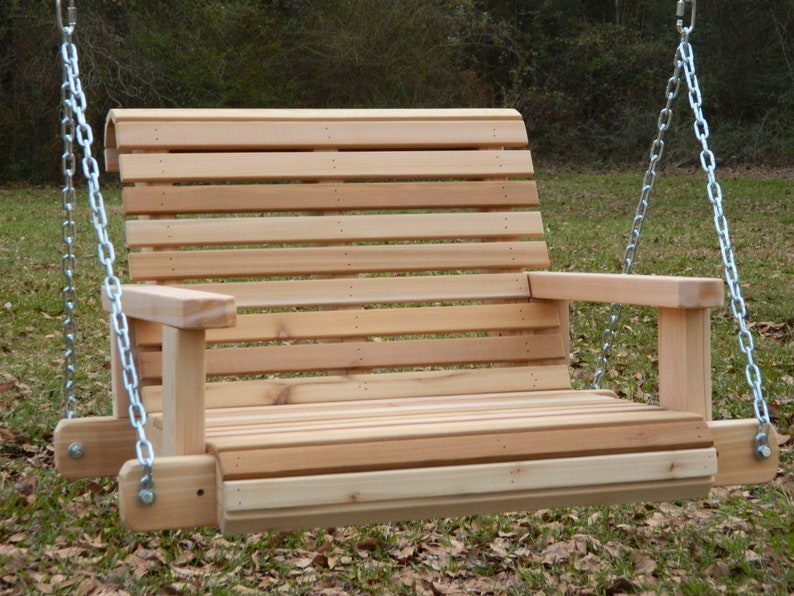 Puede incluir: Un columpio de madera con respaldo de listones y apoyabrazos, colgado de dos cadenas plateadas. El columpio est&aacute; hecho de madera de cedro natural y est&aacute; suspendido en un &aacute;rea cubierta de hierba.