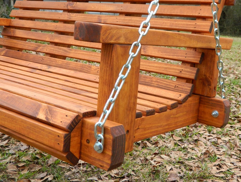 May include: Close-up of a wooden swing set with a brown stain finish. The swing is made of sturdy wood and has a metal chain hanging from it. The swing is set against a backdrop of brown leaves.