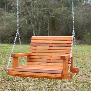 Puede incluir: Un columpio de madera con respaldo y asiento de listones, colgado de dos cadenas plateadas. El columpio est&aacute; suspendido en el aire, con un fondo de hierba y hojas ca&iacute;das.
