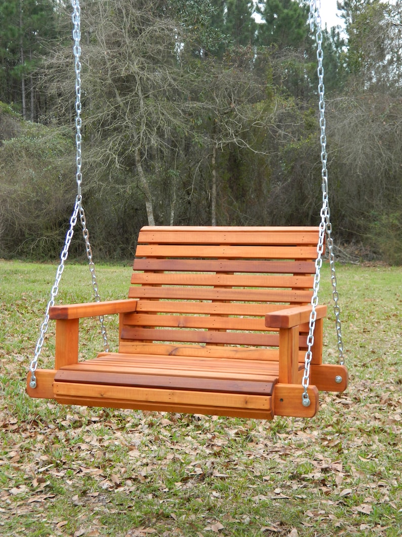 Puede incluir: Un columpio de madera con respaldo y asiento de listones, colgado de dos cadenas plateadas. El columpio est&aacute; suspendido en el aire, con un fondo de hierba y hojas ca&iacute;das.