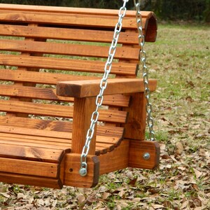 May include: A wooden swing bench with a slatted back and seat. The swing is hanging from two chains attached to the top of the bench. The bench is stained a dark brown color and is sitting on a bed of brown leaves.
