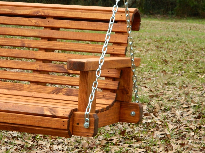 May include: A wooden swing bench with a slatted back and seat. The swing is hanging from two chains attached to the top of the bench. The bench is stained a dark brown color and is sitting on a bed of brown leaves.