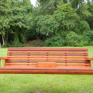 K&ouml;nnte beinhalten: Eine Holz-Schaukelbank mit einer Lattenr&uuml;cklehne und einem Sitz, die an zwei Ketten h&auml;ngt. Die Schaukel ist in einem satten Rotbraun gebeizt und &uuml;ber einem Rasenplatz aufgeh&auml;ngt.