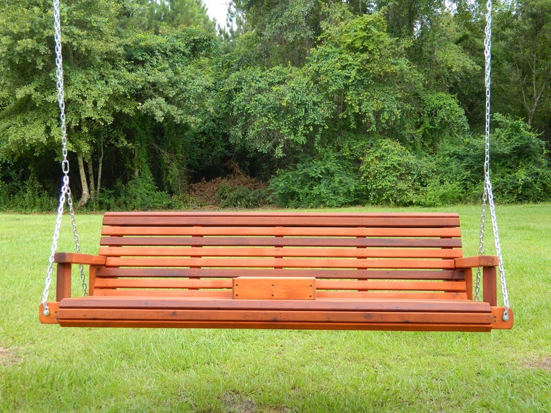 K&ouml;nnte beinhalten: Eine Holz-Schaukelbank mit einer Lattenr&uuml;cklehne und einem Sitz, die an zwei Ketten h&auml;ngt. Die Schaukel ist in einem satten Rotbraun gebeizt und &uuml;ber einem Rasenplatz aufgeh&auml;ngt.