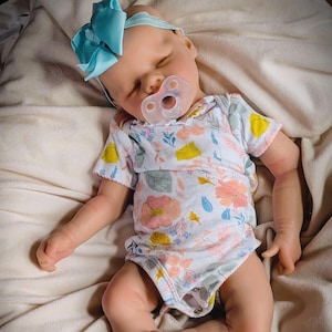 Può includere: Una bambola realistica per bambini con un body a stampa floreale e un ciuccio. La bambola ha una fascia blu con fiocco e giace su una coperta color crema. Gli occhi della bambola sono chiusi e sembra dormire.