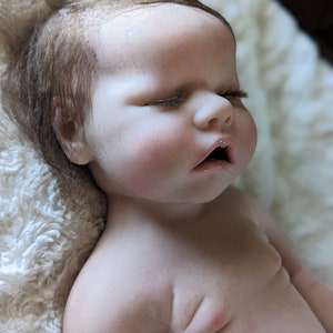 May include: A realistic baby doll with closed eyes and a slightly open mouth. The doll has brown hair and is lying on a white fluffy blanket.