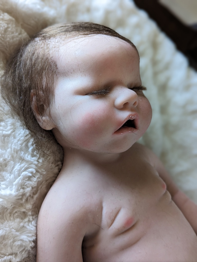 May include: A realistic baby doll with closed eyes and a slightly open mouth. The doll has brown hair and is lying on a white fluffy blanket.