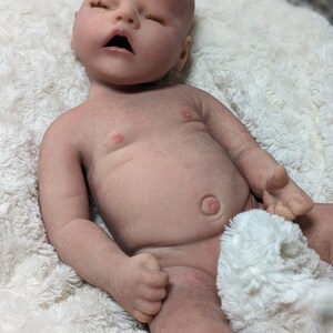 May include: A realistic baby doll made of silicone, lying on a white fluffy blanket. The doll has closed eyes, a slightly open mouth, and a belly button.