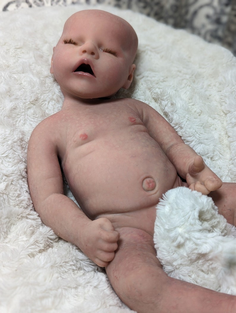 May include: A realistic baby doll made of silicone, lying on a white fluffy blanket. The doll has closed eyes, a slightly open mouth, and a belly button.