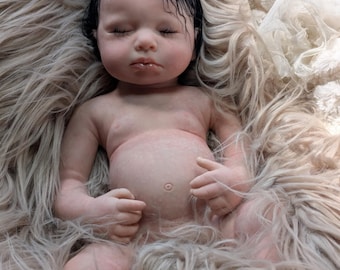 May include: A lifelike baby doll with closed eyes and dark hair, lying on a bed of fluffy, light brown faux fur. The doll has a realistic skin tone and is posed with its hands gently curled.