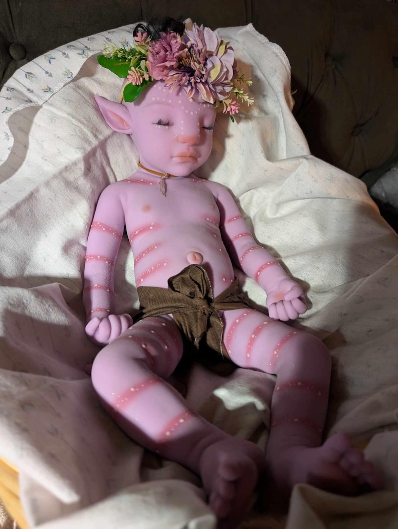 Pu&ograve; includere: Una bambola di silicone viola con strisce rosa e una corona di fiori. La bambola indossa un pannolino di stoffa marrone e giace su una coperta bianca.