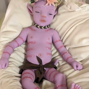 Pu&ograve; includere: Una bambola di silicone viola con una corona di fiori, che indossa un pannolino di stoffa marrone. La bambola ha dettagli dipinti, tra cui punti bianchi e strisce rosa.